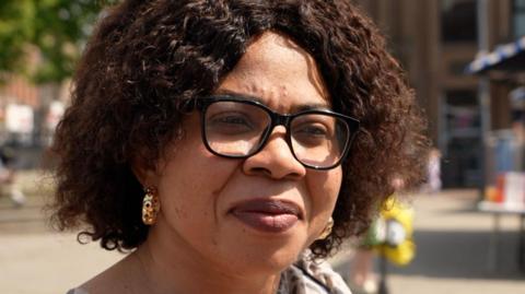 Kate Igbinigie is a black woman with curly hair cut into a bob. She's wearing black, square framed glasses and gold hoop earrings. 