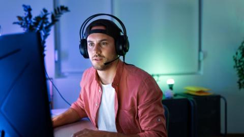 A young man in a t-shirt and open pink shirt wearing a headset with headphones and a mouthpiece is looking at a large screen with a look of concentration on his face.