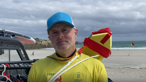 Close up of Alan Jenner at the beach. He is wearing a yellow rash vest and a blue cap. A red and yellow flag is on the beach behind him.