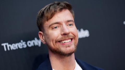 MrBeast pictured in New York in May 2025. The creator has blue eyes, short brown hair and a short beard. He smiles, wearing a dark blue blazer over a white T-shirt while posing for photographers in front of black hoarding. 