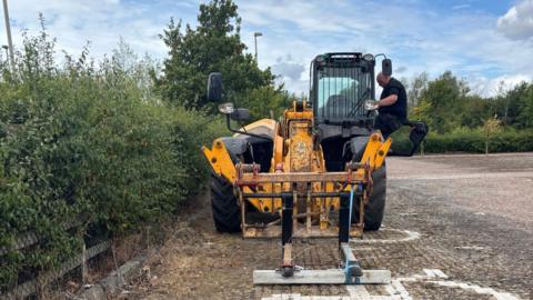 A man exiting the cab of a yellow piece of machinery, which is parked on paved car park stones beside a tall hedge. 