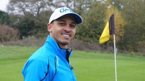 Peter Odemwingie wears a blue jacket and white cap as he stands side on and looks into the camera while standing on a golf green with a yellow and blag flag in the background to the right