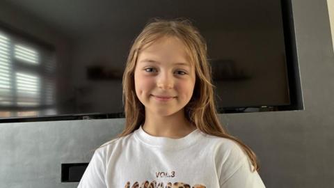 Bella Shipley stood smiling in front of a TV and a grey wall wearing a white T-shirt with White Fox logo on it.