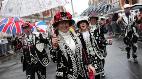 Four Pearly Kings and Queens in their distinctive, decorated clothing with umbrellas in hand take part in the New Year's Day parade. They are in the street and crowds of people can be seen in the background, standing behind barriers on the pavement