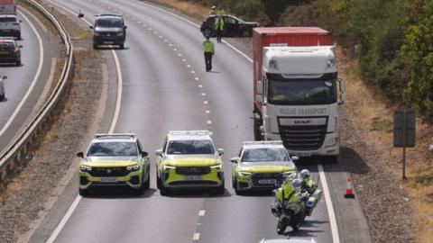 Three police cars and a motorbike blocking the dual-carriageway A12. There is a lorry with a red trailer pulled over to the side of the road. Police officers are looking at a black car in the background.