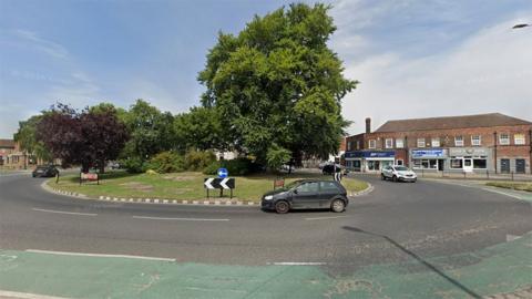 The five-road roundabout on Calvert Lane. There are a number of cars going round it and trees in the middle, with shops in the background