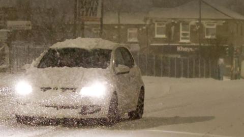 A white car, which has its headlights on, driving on a snowy street in an English village