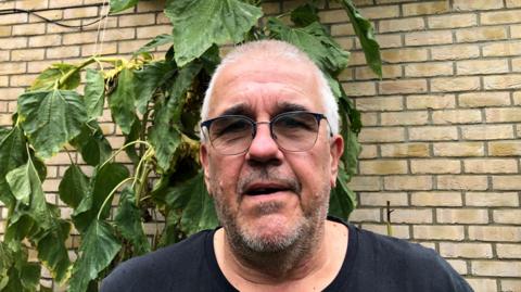 Guy Tyers, 56, with a stubbly unshaven face, short cropped grey hair and dark tinted glasses in front of part of the Fairhaven care home in Soham with his 3.6m tall sunflower, which he talks to, to encourage growth