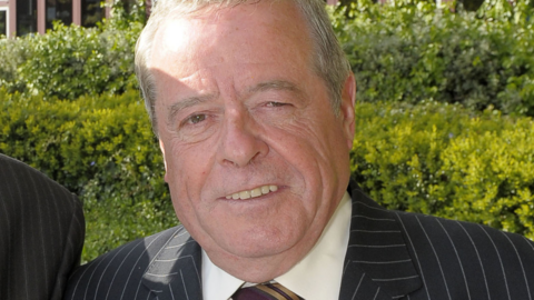 A man, who is prominent in the image and looking at the camera, is wearing a black and white striped jacket, a light shirt and a tie. Much of the background features bushes.
