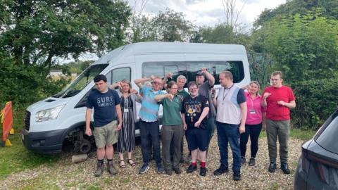 Participants of the charity next to the vandalised white minibus. Many are pointing with their thumbs down.