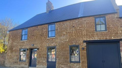The exterior of the Old Black Lion pub on a bright and sunny day. It is a brown stone building with dark grey sash style windows. There is a set of dark grey double garage doors to the right. It has a slate grey roof with a chimney stack on the top. 