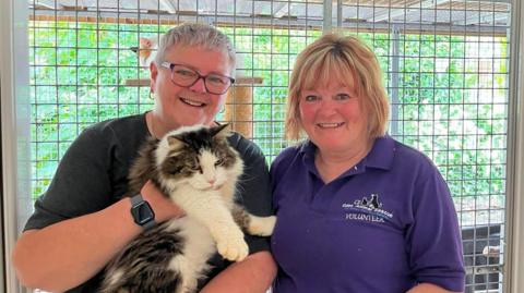 Stan is pictured in a rescue environment, being held by Elaine Brown and Sue Emmerson in front of a secure enclosure. The structured surroundings and visible signage indicate a formal animal care setting, while the close physical handling demonstrates attentive care and human interaction within the shelter. Elaine Brown has short white hair, glasses, dark top, loop earrings and a watch. Sue Emmerson has bobbed blonde hair and a purple top with a Eden Animal Rescue logo and the word "volunteer".
