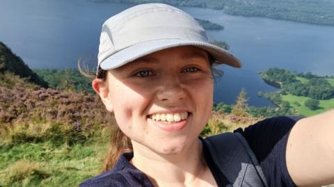  Hannah is standing high on a hill with a large lake behind her. She wears a grey baseball cap, navy top and a rucksack. She is smiling as she takes a selfie.