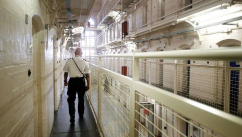 A prison officer walks away from the camera down the hallway of a prison. It's an open hallway so you can look up towards other floors. The walls are white and the fencing on the other side of the walkway towards the middle is also painted white. 