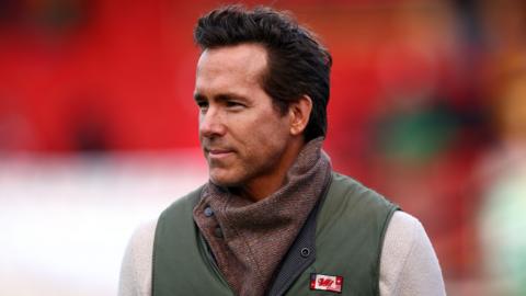 Headshot of Ryan Reynolds at the football pitch. He has dark brown hair and is wearing a white jumper with a green gilet over the top, There's a brown scarf-like item around his neck.