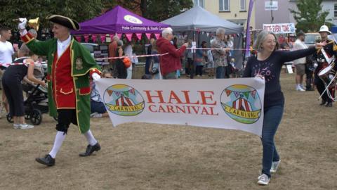 A man in a tricorn hat ringing a bell and a woman in a blue top and jeans hold a banner bearing the words "Hale Carnival"