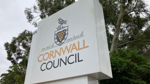 White sign reads "One and all Cornwall Council". Green trees are behind the sign. 