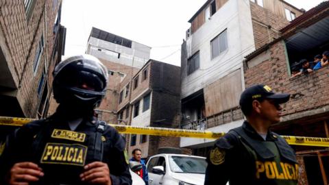 Two police officers guard a cordoned off area, which has several apartment blocks that are about four storeys high. One of them is blackened by fire.