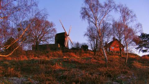 An image of the Reigate Heath Windmill, a rare Grade II listed post mill perched on the edge of the heath in Surrey. The picture has been taken from a lower vantage point, and a house can be seen to the right of the windmill. A tiny bit of snow remains on the ground. 