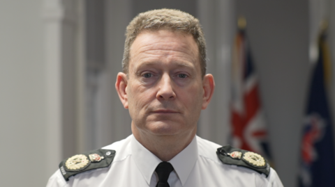 Ben-Julian Harrington is wearing a white shirt and black tie with black and gold epaulettes on his shoulders. He has short brown hair and looks at the camera with a serious expression. Behind him are a Union Jack flag and a navy Essex Police flag.