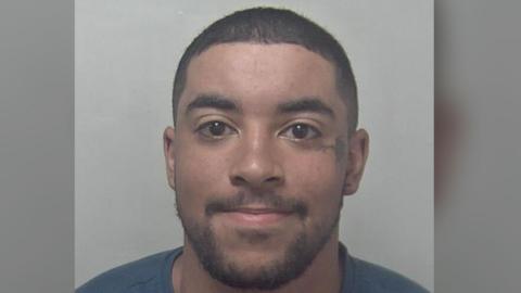 A mugshot of a man smiling and looking directly at the cameras. He is wearing a blue top. 