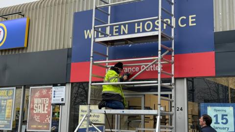 A sign outside a shop which reads - Willen Hospice, clearance outlet. There is a man with a drill on scaffolding in front of the sign. Another man is on the ground looking up. There is a sign in the window saying - opening 15 January.