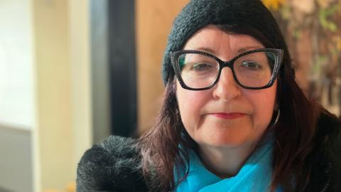 Yelena, a woman, wearing a black wooly hat, black sparkly glasses, blue scarf and a black coat, smiling for camera.