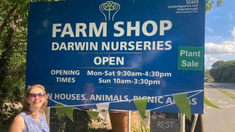 Sally wearing a blue top and sunglasses smiling - while standing next to a big blue farm shop signboard.