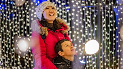 Woman wearing a red coat with a cream woolly hat stood with a boy wearing a dark coloured coat both looking up and surrounded by small white lights