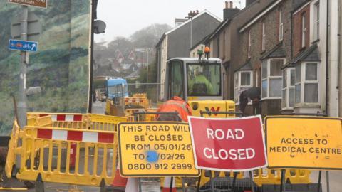 Road closure signs and barriers block off a road. Gas engineers are on the site, along with machinery. Houses are on either side of the closed road. Properties are in the distance.