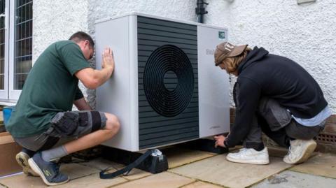 Two workers in casual clothing installing a heat pump - a large white box dominated by a black fan unit - on the outside a house with white harling walls