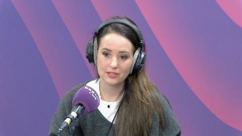 Woman with long brown hair swept to one side wearing a grey v neck sweater with a white round neck blouse underneath sat against a purple background with a BBC CWR branded microphone in front of her 