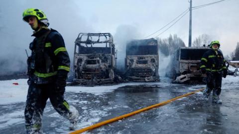 A firefighter walking in front of three burnt out semi-trucks with sleet and snow surrounding the vehicles