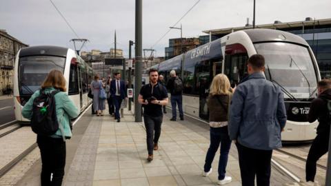 People at a tram stop in Edinburgh with two trams waiting on either side of the platform