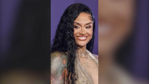 Kehlani, with long black hair and shoulder tattoos, smiles to camera, against a purple background