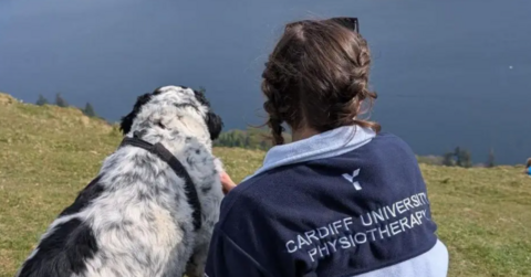 Chloe Hurst l looks out to sea while sat next to a dog. She os wearing a top which reads cardiff university physiotherapy 