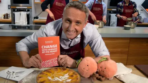 Alex Cole-Hamilton launches the manifesto at a baking school. He is holding the manifesto while leaning next to a peach tart