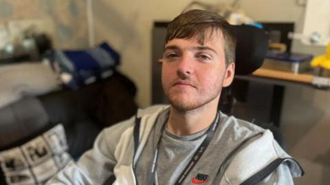  A man looks at the camera. He is wearing a tracksuit top and grey t-shirt and a lanyard. In the back is a blurry background where a seat and cushion can be seen. 