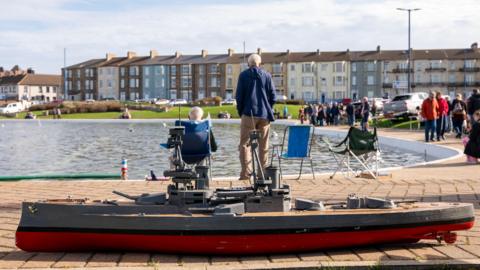 A long red and grey model boat is sitting near the boating lake in Redcar. A few smaller boats are in the water. People are walking past the lake and looking at the boats. There is a row of colourful terraced houses across the street.
