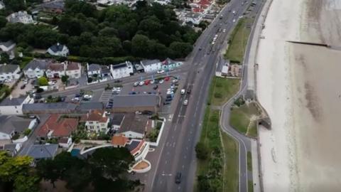 A double lane road along a stretch of sand. There is a green patch between the road and the beach and trees and buildings along the left side of the road.