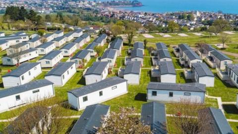 An aerial view of Beverley Park in Paignton showing static camp sites with green space, houses and the sea in the background on a sunny day.