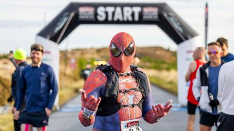 A marathon runner dressed as Spider-Man with other runners in the background looking on and the start sign for the marathon