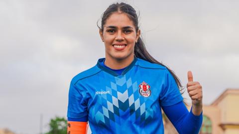 Kawtar Bentaleb, wearing a blue goalkeeper's jersey, gives a thumbs up gesture with her left hand