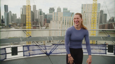 Lucie Colebeck smiling at the camera is standing on the roof of the O2. She is wearing black trousers and a purple top.