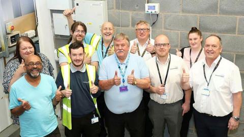 A group of 10 people, including men and women, some in hi-viz, smile up at a camera and give the thumbs up