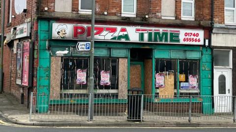 A badly burnt and smashed building with the name Pizza Time displayed on a board above the entrance. A bin and a metal fence are on the pavement outside.