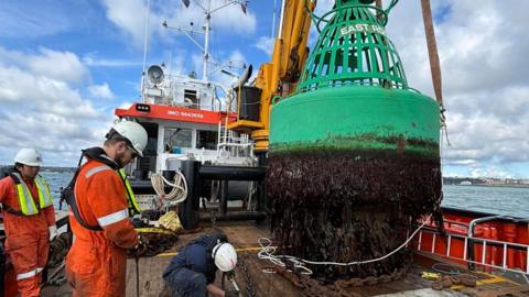 The East Rock buoy onboard the Bommel tugboat. Seaweed is covering the bottom of the large green buoy. Two workers dressed in orange are watching as a workman, dressed in navy, inspects the buoy's chain. 