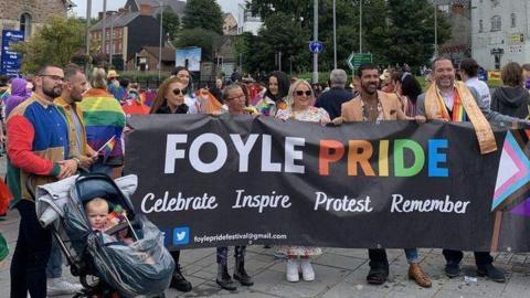 A group of people hold a foyle pride banner as they stand outside Derry's railway station, with a large crowd behind them. A number of buildings can be seen in the background while a man and a baby in a pram are standing to their right
