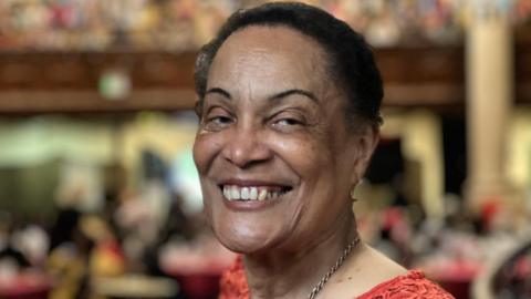 Antonette, a lady in her 80s, smiles in a busy room, which is blurred behind her. She is wearing a gold necklace and a red top, and her black hair is back.