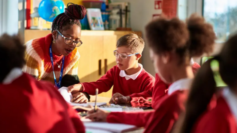 A teacher helps a pupil in a classroom with other pupils sat by him at desks. The teacher and male pupil both have glasses on. The children are wearing a red school uniform.